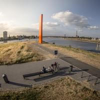 Orange Rheinorange sculpture stands prominently on grassy hill overlooking Rhine River in Duisburg with cyclists on viewing platform below.