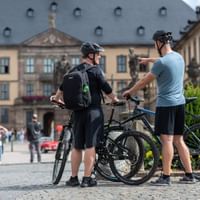 Two cyclists with helmets and bikes pause in Fulda's historic city center, with baroque buildings and cobblestone square in background.