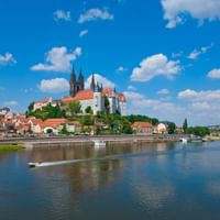 Historische Silhouette von Meißen mit gotischem Dom und Burg auf dem Hügel über der Elbe. Rotgedeckte Gebäude säumen das Flussufer unter blauem Himmel.