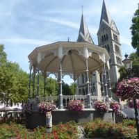 Weißer viktorianischer Musikpavillon mit verzierten Säulen im Park am Dom, Roermond, mit zweitürmiger Kathedrale dahinter und bunten Blumenbeeten.
