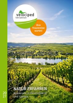 Velociped Katalog 2027 mit Weinbergreihen, die zu einem Flusstal mit Dorf, Hügeln und Windrädern unter blauem Himmel führen.
