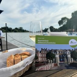 Münsterland Schlösserführer auf Holztisch mit Weinglas und Brotkorb, mit Blick auf einen See mit Booten und Bäumen im Hintergrund.