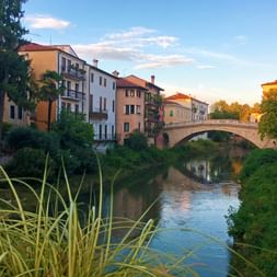 Stone bridge spanning a canal in Vicenza with colorful historic buildings lining the waterway. Lush vegetation grows along the banks under a blue sky.