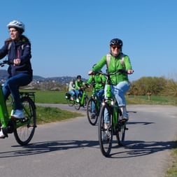 Gruppe von Radfahrern auf grünen E-Bikes auf einem asphaltierten Radweg bei Marburg. Blauer Himmel, grüne Felder und Hügel im Hintergrund.