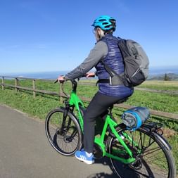 Radfahrer mit türkisfarbenem Helm auf grünem E-Bike entlang des Fulda-Radwegs mit Blick auf Hügel und ferne Berge unter blauem Himmel.