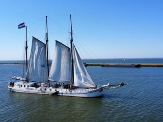 Zweimaster-Segelschiff Elizabeth mit weißen Segeln auf ruhigem blauen Wasser unter klarem Himmel, mit Küstenlandschaft im Hintergrund.