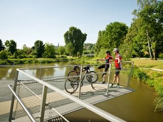 Zwei Radfahrer mit Fahrrädern stehen auf einer modernen Metallbrücke über einem Fluss im Bamberger Erba Park, umgeben von grünen Bäumen.