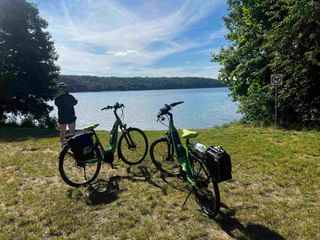 Two e-bikes with panniers parked on grass at Werbellinsee bathing spot. A person stands at the shore viewing the lake and forested hills.
