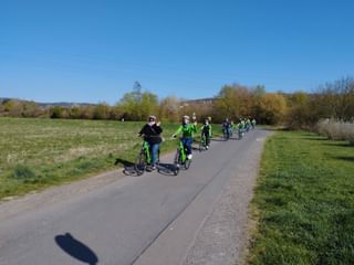 Gruppe von Radfahrern auf asphaltiertem Weg durch grüne Wiesen unter blauem Himmel. Bäume säumen den Weg, Hügel in der Ferne.