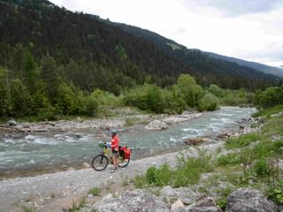 Radfahrer in roter Jacke mit Tourenrad am steinigen Ufer der türkisfarbenen Drau, bewaldete Berge im Hintergrund unter bewölktem Himmel.