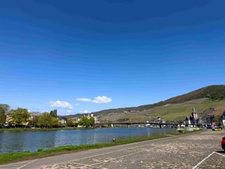 Panoramablick auf Bernkastel-Kues an der Mosel mit Kirchturm, Brücke, Booten und weinbergbedeckten Hügeln unter blauem Himmel.