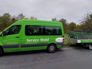 Green van labeled 'Service Mobil' pulling a trailer loaded with multiple bicycles, parked on a road with trees in the background.