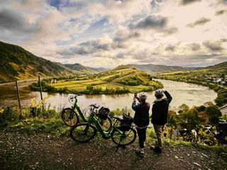 Two cyclists with bikes viewing the Mosel river loop from a hilltop. The river curves around a green peninsula with vineyards and hills in the background.