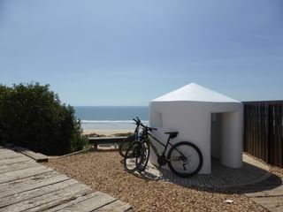 Zwei Fahrräder neben einem weißen zylindrischen Strandunterstand auf Kiesboden, mit Meer und Sandstrand im Hintergrund.