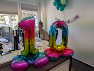 Frau steht am Büroschreibtisch mit großen regenbogenfarbenen Zahlenballons 10, Girlande und Computermonitoren in hellem Arbeitsbereich.