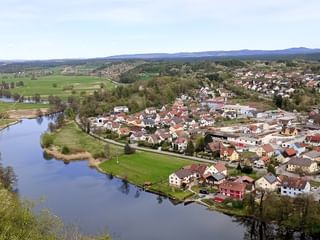 Panoramablick auf das Dorf Kallmünz mit rotgedeckten Häusern entlang eines Flusses, umgeben von grünen Wiesen und fernen Hügeln unter blauem Himmel.