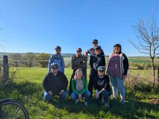 Gruppe von neun Radfahrern posiert in zwei Reihen auf einem grasbewachsenen Hang mit grünen Feldern und blauem Himmel im Hintergrund.