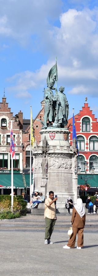Historischer Grote Markt Platz in Brügge mit bunten mittelalterlichen Gebäuden, zentralem Denkmal und Touristen auf dem Platz.