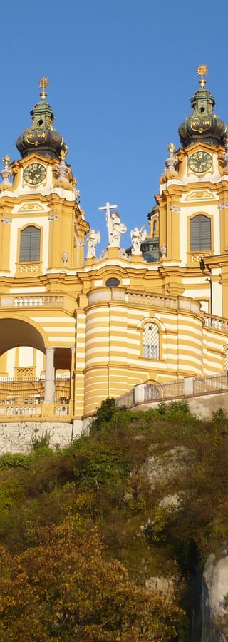 Gelbes Barockkloster mit Zwillingstürmen und grünen Kuppeln thront auf einem bewachsenen Felsenhügel vor klarem blauen Himmel.