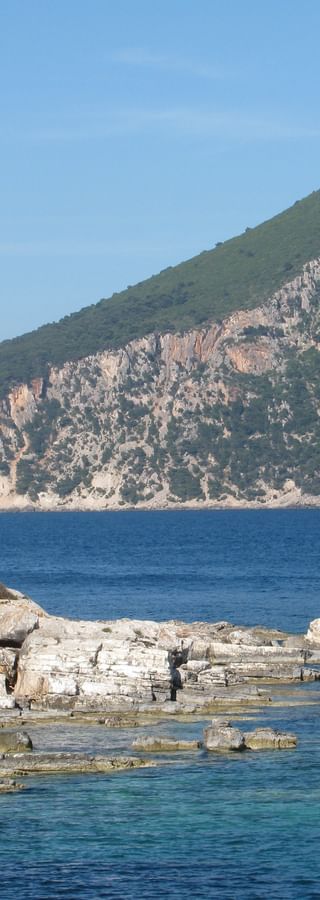Steinerner Wachturm an felsiger Küste der Ionischen Inseln mit blauem Meer und bergiger Landschaft im Hintergrund unter klarem Himmel.