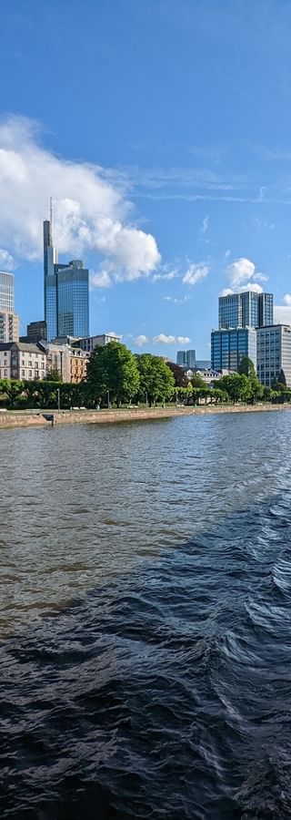 Blick auf Frankfurter Skyline mit Wolkenkratzern vom Bootsdeck auf dem Main. Blauer Himmel mit weißen Wolken über der Uferpromenade.