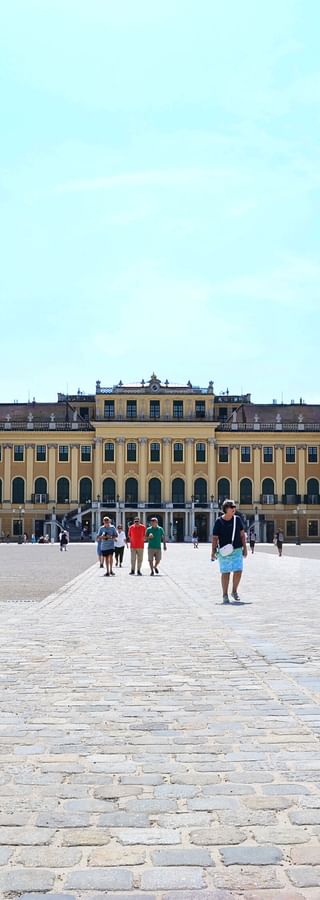 Gelbes barockes Schloss Schönbrunn in Wien mit seinem großen Ehrenhof. Besucher gehen über den gepflasterten Platz unter blauem Himmel.