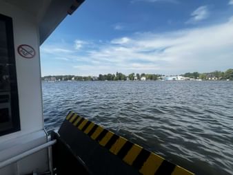 Blick vom Fährdeck mit gelb-schwarz gestreifter Rampe über Wasser zum bewaldeten Ufer bei Grünau. Blauer Himmel mit vereinzelten Wolken.