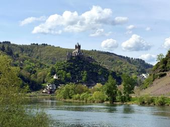 Reichsburg Cochem thront auf einem bewaldeten Hügel über der Mosel, mit Weinbergen am rechten Ufer und weißen Wolken darüber.