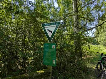 Green triangular sign reading 'Naturschutzgebiet' with bird silhouette, mounted on post with information board in sunny forest. Bicycle visible at right.