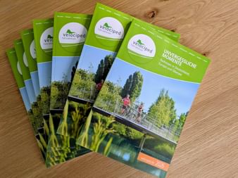 Stack of green Velociped cycling tour catalogs on wooden surface, featuring cyclists on a bridge over water with trees in background.