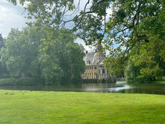 Wasserschloss Darßfeld umgeben von Wasser und üppigen grünen Bäumen, mit gepflegtem Rasen im Vordergrund unter teilweise bewölktem Himmel.