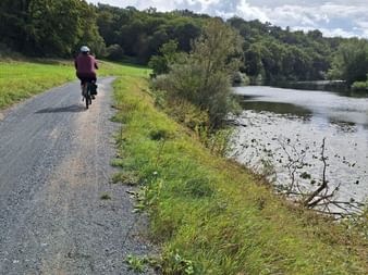 Radfahrer auf asphaltiertem Weg entlang eines Flusses auf dem Radweg Wetzlar Weilburg. Grüne Wiesen und bewaldete Hügel unter bewölktem Himmel.