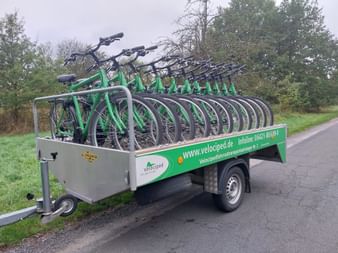Green Velociped bike trailer loaded with multiple green bicycles parked on a road beside grass and trees.