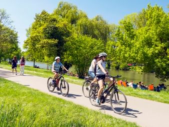 Two cyclists with helmets riding on paved path along Neckar River, surrounded by lush green trees and grass with people relaxing by water.