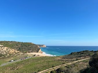 Panoramablick auf den Boca do Rio Strand an der Algarve, Portugal mit goldenem Sandstrand, türkisfarbenem Meer, grünen Hügeln und blauem Himmel.