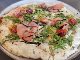 Dünnkrustige Pizza belegt mit geräuchertem Lachs, frischem Rucola, Kirschtomaten und Balsamico-Glasur auf einem weißen Teller.