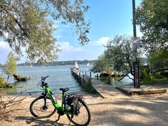 Grünes E-Bike am Radweg neben dem Wannsee geparkt. Holzsteg führt ins Wasser, Segelboote am Horizont unter blauem Himmel sichtbar.
