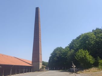 Hoher Ziegelschornstein der Zeche Nachtigall mit rotgedecktem Gebäude und Wald unter klarem blauen Himmel im Ruhrgebiet.