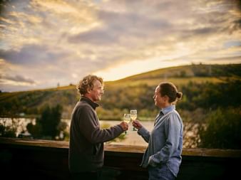 Man and woman toasting with white wine glasses at sunset overlooking Mosel River and vineyard hills during golden hour.