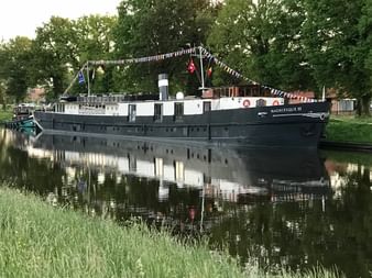 Flusskreuzfahrtschiff MS Magnifique III mit dunklem Rumpf und weißem Aufbau am ruhigen Gewässer, geschmückt mit bunten Flaggen.