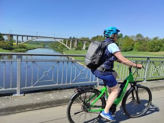 Radfahrer mit blauem Helm und grünem Fahrrad auf Brücke über der Fulda. Große Bogenbrücke aus Beton im Hintergrund unter blauem Himmel.