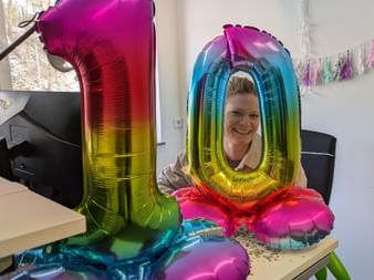 Smiling woman named Carolin at desk with rainbow-colored number 10 balloons celebrating anniversary in office with colorful decorations.