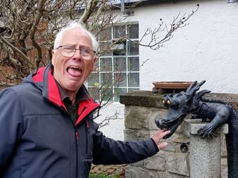 Mann in rot-schwarzer Jacke posiert spielerisch mit einer dunklen Metall-Drachenbrunnen-Skulptur in Dietfurt, weißes Gebäude im Hintergrund.