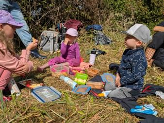 Four children sitting on straw having a picnic with lunchboxes and backpacks at an outdoor event in Burgwald forest.