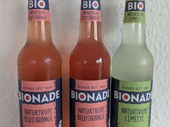 Three Bionade bottles: two blood orange and one lime flavored isotonic drinks with dark caps, standing against a white textured wall.
