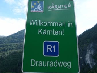 Grünes Schild mit 'Willkommen in Kärnten' und 'Drauradweg R1' mit Radrouten-Symbolen vor bewaldeten Bergen und blauem Himmel.