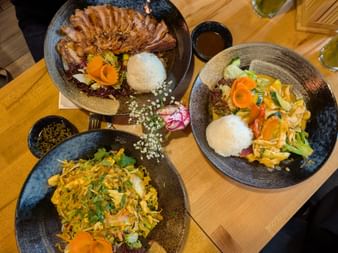 Drei asiatische Gerichte auf dunklen Keramiktellern mit weißem Reis, Entenscheiben, gebratenen Nudeln und buntem Gemüse auf einem Holztisch.