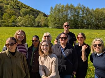 Group of eight people posing in a yellow flowering meadow with trees and hills in the background under blue sky.