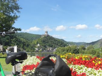 Grünes Fahrrad neben buntem Blumenbeet am Moselradweg bei Cochem, mit Reichsburg auf dem Hügel im Hintergrund sichtbar.