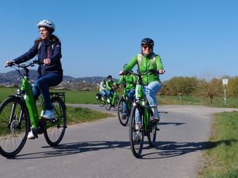 Group of cyclists on green e-bikes riding on a paved path through countryside with hills in the background under clear blue sky.